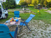 Tisch mit Tellern, Bechern und Flaschen; drei blaue Klappstühle; weißes Camperfahrzeug links; Wiese mit erdigen Stellen; Holzsteg und überdachte Plattform über Wasser; Bäume im Hintergrund; gelbes Schild