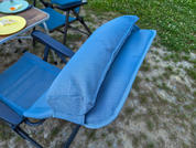 Blauer gepolsterter Aufsatz auf der Armlehne eines blauen Klappstuhls; im Hintergrund ein Tisch mit Tellern und Grasboden