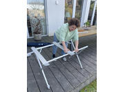 Una persona se inclina sobre una mesa plegable blanca doblada en una terraza de madera frente a una puerta corredera de vidrio