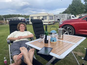 Persona en una silla de camping con los pies apoyados en una mesa plegable; en la mesa una botella de vino, una copa de vino, una linterna azul, una jarra metálica y una tableta; al fondo caravanas, coches, césped y árboles bajo un cielo nublado