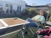 Persona sentada en una silla plegable en el jardín, viste prenda verde claro y vaqueros; mesa con superficie de madera y un smartphone; casa blanca con puertas de terraza al fondo; parte de una segunda persona en primer plano