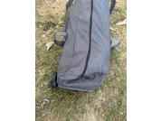 Sac gris avec fermeture éclair et roulette visible posé sur de l'herbe et de la terre, feuilles sèches à côté