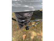 Compartiment en tissu gris avec plusieurs poches fixé à une structure métallique noire avec petites roues noires ; protection verte de roue ; sol avec herbe et feuilles sèches