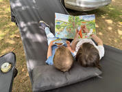 Deux enfants allongés côte à côte sur un hamac gris tenant un livre ouvert avec des illustrations colorées ; l'un tient un petit bol de snacks. En arrière-plan herbe, ombres et une voiture garée