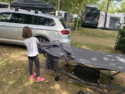 Enfant de dos en t-shirt blanc et chaussures roses debout à côté d'un lit de camp plié devant une voiture break argentée avec coffre de toit sur un camping ; camping-cars et arbres en arrière-plan