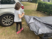 Personne aux cheveux châtain mi‑longs, portant un T‑shirt blanc, des leggings noirs et des chaussures roses, debout sur l'herbe à côté d'une voiture argentée et tenant un coin d'une bâche grise sur une remorque, buissons en arrière‑plan