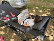 Un enfant est allongé sur une couchette de camping noire avec un coussin et tient un livre d'images coloré ouvert; porte un t-shirt blanc, un legging gris et des chaussettes roses; en arrière-plan une voiture argentée et des arbres avec des ombres; une chaussure rose au sol