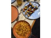 Tisch mit zwei Spießen mit braunen Stücken und grünen sowie roten Gemüsescheiben auf einem hellblauen Teller; eine orange Schüssel mit gewellten Pommes; eine Schüssel mit oranger Suppe auf einem rosa Teller; ein Glas mit bernsteinfarbener Flüssigkeit