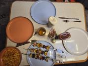 Tisch mit vier Tellern, Schale mit Spiralnudeln, Teller mit zwei Metallspießen mit Fleisch- und Gemüsestücken, Glas mit Getränk, Flasche mit brauner Sauce, kleines Schälchen mit grüner Paste und Besteck