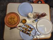 Tisch mit zwei blauen Tellern, einem weißen Teller, einer Schale Pommes, zwei Metallspießen mit Stücken aus Gemüse und Fleisch, zwei Saucenflaschen, einem Salzstreuer und einem kleinen Glas