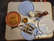 Aufsicht auf einen Tisch mit vier Tellern: links Terrakotta-Schüssel mit gewellten Pommes, unten blauer Teller mit zwei Metallspießen mit gelb-braunen Stücken, rechts leerer weißer Teller, oben leerer blauer Teller; in der Mitte kleines Glas mit gelb