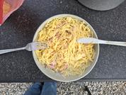 Schüssel mit gelben Nudeln in heller Sauce und kleinen rosa Stücken; Gabel und Messer in der Schüssel; dunkle Arbeitsplatte; grauer Topf oben rechts; pinke Verpackung oben links; Hosenbeine und Kiesboden am unteren Bildrand