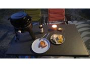 Tisch im Abendlicht mit zwei Tellern (links ein Stück Pizza mit Besteck, rechts mehrere gelbe Scheiben und eine hellere Stelle), brennende Kerze, Flasche, kleines rechteckiges Packstück, Grill links, zwei Klappstühle (grün, orange), Kiesboden und gem