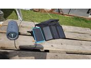 Zylindrischer Lautsprecher, Smartphone mit blauem Rahmen und ein aufgeklapptes dreiteiliges Solarpanel mit sichtbaren Kabeln liegen auf einer Holzpalette; im Hintergrund Rasen und eine Jacke