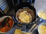 Korb einer Heißluftfritteuse mit geriebenem Käse auf Lebensmitteln, daneben Schalen mit weiteren Zutaten auf einer Arbeitsfläche