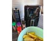 Black air fryer on a countertop, in front a green bowl with sliced bananas, to the left several spice jars and a tall dark bottle, white blinds in the background