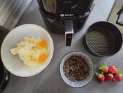 Black air fryer with 'Berger' logo on a countertop; white bowl with two cracked eggs (visible yolks and white pieces); small bowl with dark brown crumbly contents; round black perforated basket; three strawberries