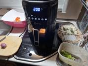 Friggitrice ad aria nera con display blu "200 °C" su lavello in acciaio inox, accanto a un tagliere con scalogno, una ciotola rosa con pezzi arancioni, confezione di pane piatto e contenitore di plastica con lattuga verde