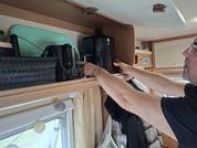 Persona indica con entrambe le mani un pensile aperto in un camper; dentro ci sono due dispositivi neri e una borsa nera intrecciata; sotto una finestra con tenda e lucine a catena