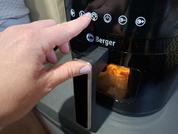 Mano preme un pulsante su un apparecchio nero con il logo "Berger", icone di controllo illuminate e un vano estraibile con finestra che mostra cibo di colore arancione