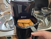 Apparecchio da cucina nero con la scritta 'Berger', cassetto aperto con diversi panini marroni; mano destra che tiene la maniglia; sullo sfondo lavello, filtro per caffè con filtro e pentola sul fornello