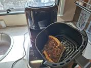 Friggitrice ad aria sul piano cucina, cestello aperto con una fetta di pane con copertura scura e pezzetti verdi, lavello e finestra sullo sfondo