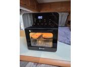 Forno da tavolo compatto nero con display digitale 180; due fette di pane su una teglia all'interno; mobili in legno, una tenda e una coperta viola sullo sfondo