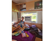 Person sitzt auf einem Bett in einem Wohnwagen; auf dem Bett liegen Kuscheltiere, Bücher und Decken; an der Wand ein Fenster mit Klimagerät und Oberschränke