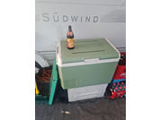 Grüne Kühlbox mit einer Flasche auf dem Deckel; rechts eine rote Getränkekiste; links ein aufgewickeltes oranges Kabel; Rückwand mit der Aufschrift SÜDWIND