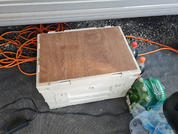 Weißer rechteckiger Kunststoffkasten mit braunem Holzbrett oben, orangefarbene Kabel im Hintergrund, rechts grüne Verpackung mit Flaschen und mehrere Plastikflaschen, links schwarzes Netzteil und Kabel auf grauem Boden vor einer weißen Wand mit horiz