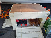 Offene beige Kunststoffkiste mit Holzbrett oben; innen drei Kunststoffflaschen mit roten Verschlüssen und dunkler Flüssigkeit sowie weitere Flaschen mit orangefarbenen Verschlüssen; links aufgerolltes orange Kabel und schwarzes Netzteil; rechts grüne