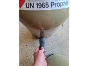 Main tenant une poignée d'allumage noire avec bouton rouge devant une bouteille de gaz verte étiquetée "UN 1965 Propan" sur un tapis beige ; une deuxième bouteille est posée à côté