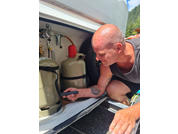 Personne chauve agenouillée devant un compartiment ouvert d'un véhicule contenant deux bouteilles de gaz, tenant un appareil noir dans une main ; tatouage sur l'avant-bras, porte un haut sans manches et un short, pneu, arbres et ciel bleu en arrière-plan