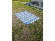 Rectangular blanket with blue, white and green zigzag stripes lies on grassy, gravelly ground; in the background a building facade with windows and vents