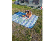 Person sitting on a blue-and-white patterned picnic blanket in front of a parked caravan; four plates and a bowl of fruit on the blanket; surrounded by grass and gravel