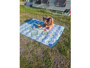 Person sitting on a blue-and-white zigzag picnic blanket on grass and gravel next to a parked caravan; on the blanket a bowl of fruit, three green plates and a pink cup