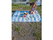 A person sits on a blue-and-white chevron blanket drinking from a cup. On the blanket are three plates, a bowl of fruit and a pitcher. In the background a camper van and a white tent. Ground of grass and gravel