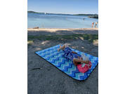 Person lying on a blue-and-white chevron beach mat on the sand, holding a book over their face; pink bag beside them, sea with a few people in the water and trees on the horizon