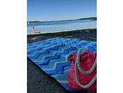 Blue and white zigzag beach mat in the foreground; pink bag with white rope handles on the mat; pebbly shore; water with shoreline and a person standing at the water; blue sky