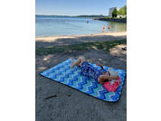 Person lies on a blue zigzag beach mat on the sandy shore of a lake, wearing a blue floral dress and holding a white hat; pink bag beside the mat; two people at the water and a building on the right