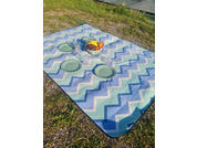 Blue-and-white zigzag blanket on grass, four pale green plates, four clear cups and a clear bowl with bananas and red and dark fruits