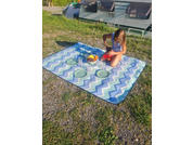 A child sits on a blue-and-white chevron blanket on grass, holding a red cup; four green plates and a bowl of fruit are on the blanket; a caravan is visible in the background