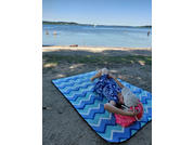 Person lying on a blue-and-white zigzag beach mat on sand, wearing a blue floral dress and white cap, holding a pink bag; water in the background with swimmers and a sailboat on the horizon
