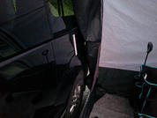 Schwarze Fahrzeugseite mit sichtbarem Rad neben einer hellen Zeltwand; schmaler Spalt zwischen Fahrzeug und Zelt, Zeltboden und ein zusammengeklappter Campingstuhl mit blauen Akzenten sichtbar