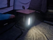 Innenraum mit hellen Stoffwänden, kleiner quadratischer Klapptisch mit rundem Objekt darauf, darunter ein schwarzer Kasten mit hellem senkrechtem Lichtstreifen, karierter Bodenmatte, Klappstuhl und ein Arm in blauer Jacke am linken Bildrand