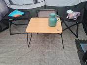 Binnenkant van een tent met twee opvouwbare campingstoelen en een lage houten tafel ertussen met een groene thermosfles; blauwe en oranje textiel op de linkerstoel, een dienblad met servies en bestek op de rechterstoel, grijze vloermat en een zwart puzzelmatstuk aan de rechterrand