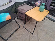 Opvouwbare houten tafel met een groene drinkfles; daarnaast een opvouwbare stoel met een kleurrijk kussen en een grijze stoffen opbergdoos op een geruit vloerkleed, tentwanden zichtbaar