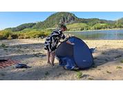 Person baut ein kleines Pop-up-Zelt am sandigen Flussufer auf, im Hintergrund Wasser und bewachsene Hügel