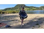 Person in schwarz-weiß gemustertem Kleid und Sonnenbrille hält einen aufgeblasenen Schwimmring auf einem sandigen Flussufer; Decke und Sandalen auf dem Sand im Vordergrund; Fluss und grüne Hügel im Hintergrund