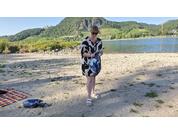 Person in schwarz-weiß gemustertem Kleid und Sonnenbrille hält eine blaue Tasche, steht auf sandigem Flussufer; links Decke und Schuhe, im Hintergrund Fluss, grüne Hügel und blauer Himmel